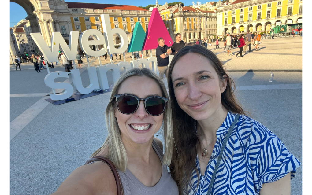 A selfie of Miriam and Hope outdoors in front of a "Web Summit" sign. The plaza features bright yellow buildings and an archway, with people gathered in the background.