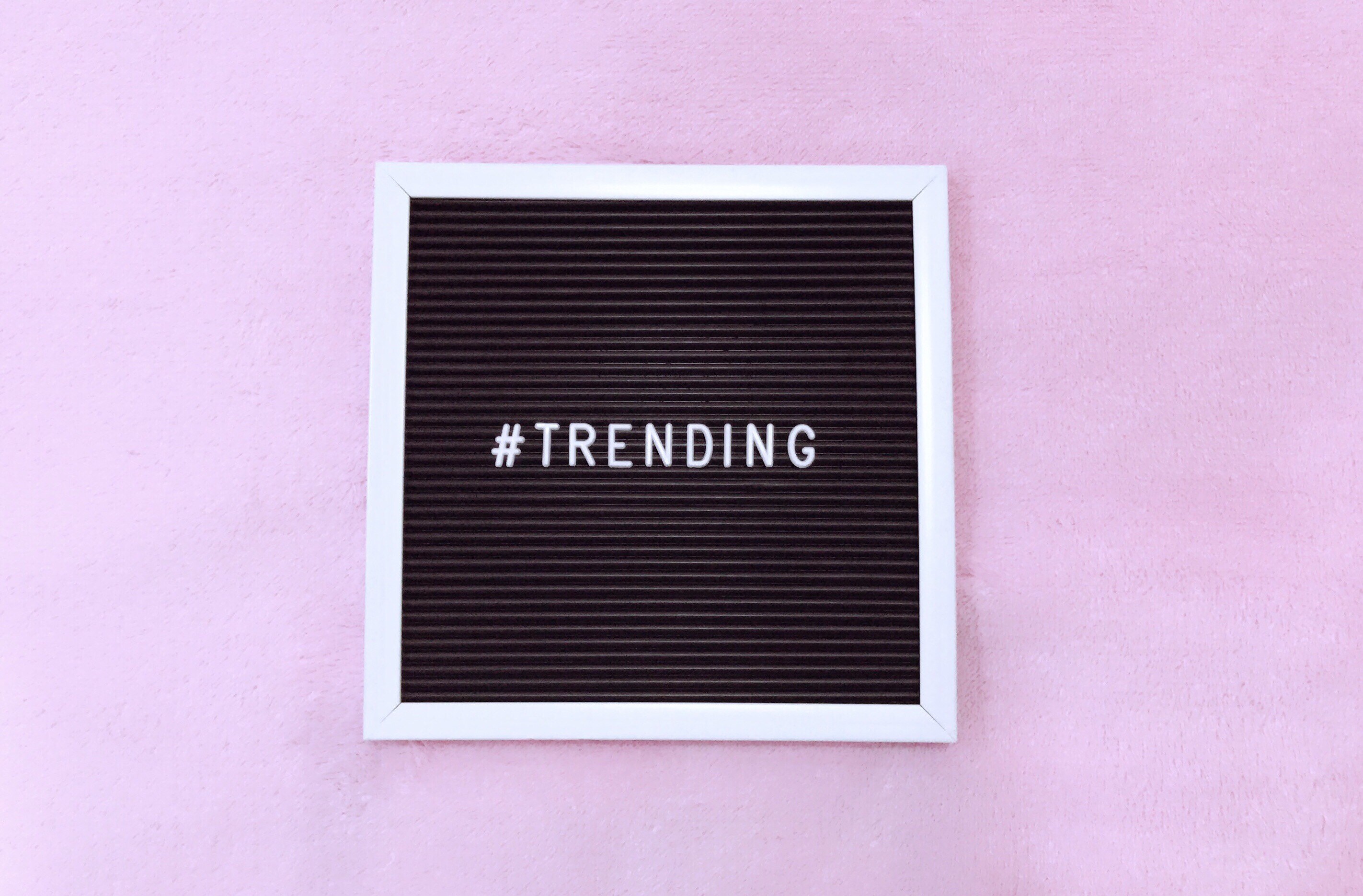 A close-up of a black message board with white letters spelling "#TRENDING". The board has a ribbed texture and a white frame, set against a soft pink backdrop.